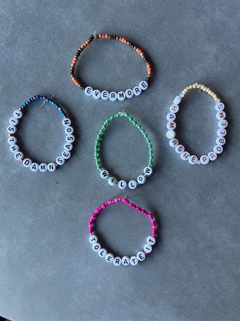 May include: Five beaded bracelets featuring white letter beads spelling out EVERMORE, TIS THE DAMN SEASON, WILLOW, CHAMPAGNE PROBLEMS, and TOLERATE IT. Each bracelet uses different colored seed beads including brown, blue, green, cream, and pink.