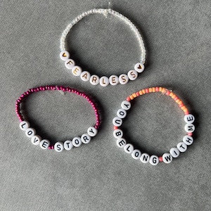 May include: Three circular beaded bracelets. The top bracelet features clear beads and white letter beads spelling FEARLESS with gold heart accents. The bottom left bracelet has magenta beads and white letter beads spelling LOVE STORY. The bottom right bracelet has orange and pink beads and white letter beads spelling YOU BELONG WITH ME.