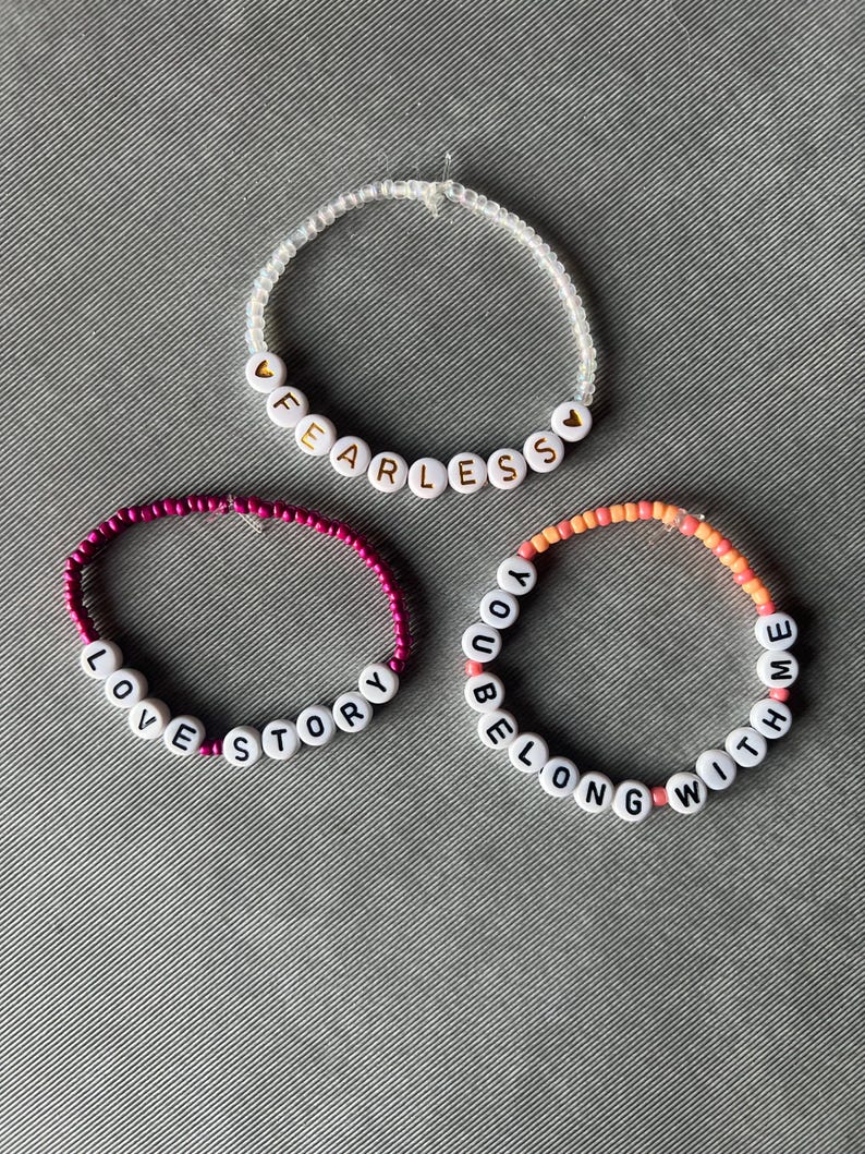 May include: Three circular beaded bracelets. The top bracelet features clear beads and white letter beads spelling FEARLESS with gold heart accents. The bottom left bracelet has magenta beads and white letter beads spelling LOVE STORY. The bottom right bracelet has orange and pink beads and white letter beads spelling YOU BELONG WITH ME.