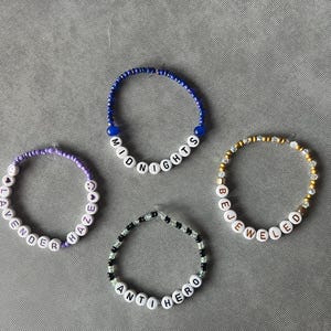 May include: Four circular beaded bracelets featuring white letter beads spelling out MIDNIGHTS, LAVENDER HAZE, BEJEWELED, and ANTI HERO. The bracelets are constructed with various colored beads including blue, purple, gold, and black and white patterns.