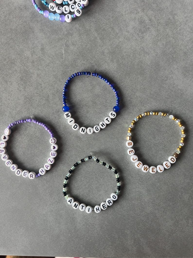 May include: Four circular beaded bracelets featuring white letter beads spelling out MIDNIGHTS, LAVENDER HAZE, BEJEWELED, and ANTI HERO. The bracelets are constructed with various colored beads including blue, purple, gold, and black and white patterns.