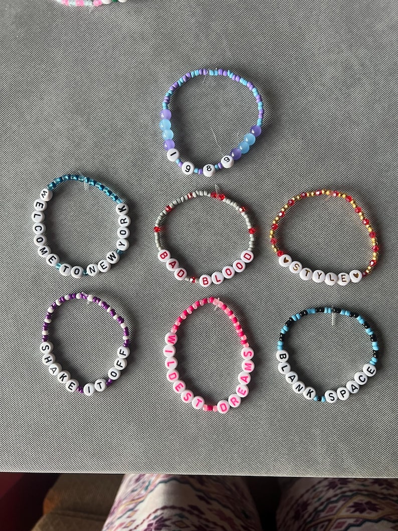 May include: Seven circular beaded bracelets featuring colorful plastic beads and white letter beads. The bracelets display text including 1989, Welcome to New York, Bad Blood, Style, Shake It Off, Wildest Dreams, and Blank Space with heart accents.