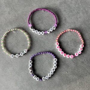 May include: Four beaded bracelets featuring white letter beads spelling out "SPEAK NOW", "OURS", "SPARKS FLY", and "BACK TO DECEMBER". The bracelets are constructed from small colorful glass beads in shades of purple, green, pink, and yellow, with some translucent and opaque finishes.