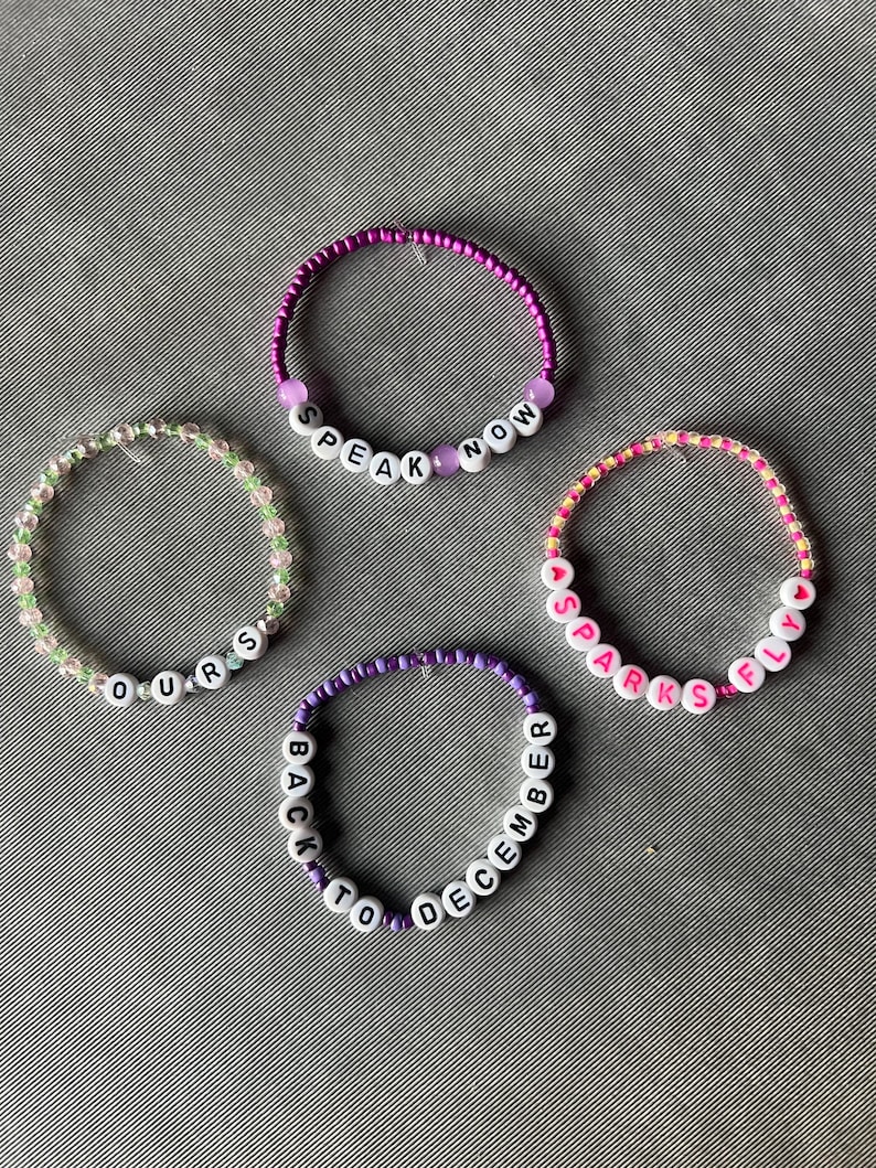 May include: Four beaded bracelets featuring white letter beads spelling out "SPEAK NOW", "OURS", "SPARKS FLY", and "BACK TO DECEMBER". The bracelets are constructed from small colorful glass beads in shades of purple, green, pink, and yellow, with some translucent and opaque finishes.
