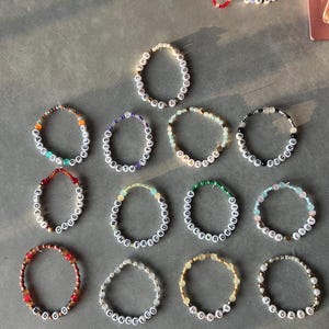 May include: A collection of circular beaded bracelets featuring white letter beads spelling phrases such as THE LIFE OF A SHOWGIRL, FATE OF OPHELIA, ELIZABETH TAYLOR, OPALITE, FATHER FIGURE, ELDEST DAUGHTER, RUIN THE FRIENDSHIP, ACTUALLY ROMANTIC, WISH LIST, WOOD, CANCELED, HONEY, and LOASG.