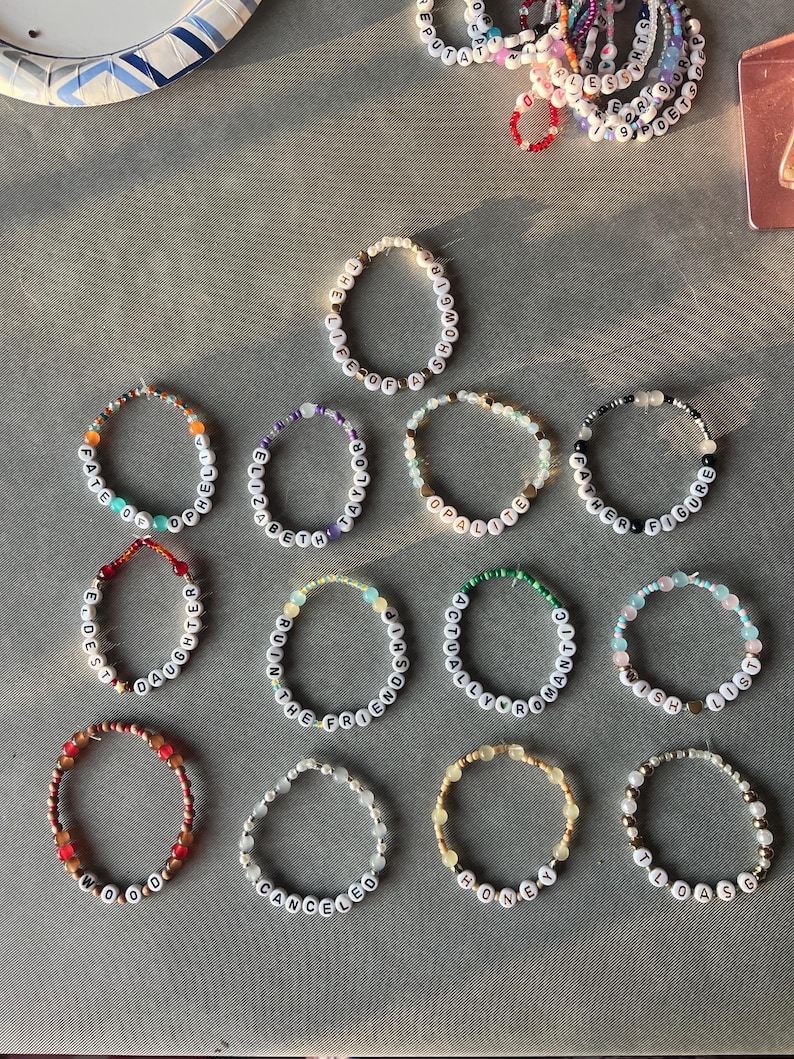May include: A collection of circular beaded bracelets featuring white letter beads spelling phrases such as THE LIFE OF A SHOWGIRL, FATE OF OPHELIA, ELIZABETH TAYLOR, OPALITE, FATHER FIGURE, ELDEST DAUGHTER, RUIN THE FRIENDSHIP, ACTUALLY ROMANTIC, WISH LIST, WOOD, CANCELED, HONEY, and LOASG.