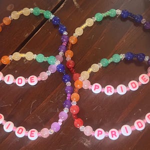 May include: Two circular beaded bracelets featuring a rainbow color sequence of red, orange, yellow, green, blue, and purple beads. Each bracelet includes white round beads with pink lettering that spell out the word PRIDE in a repeating pattern.