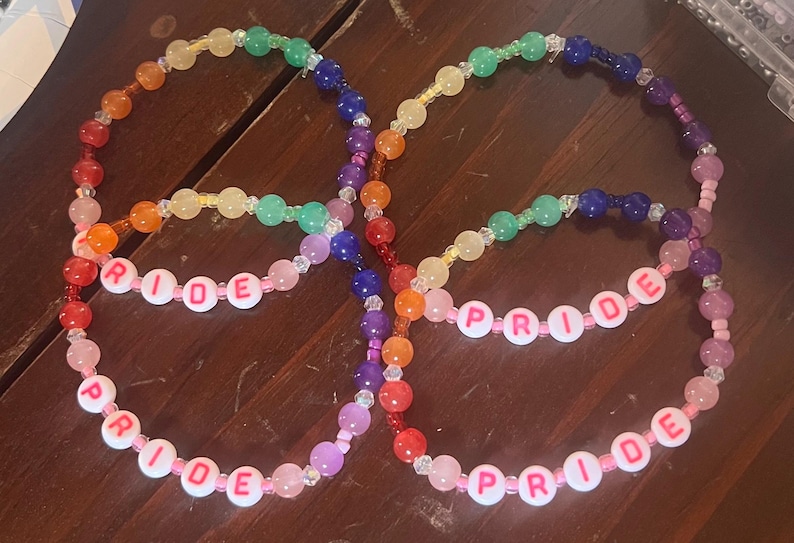 May include: Two circular beaded bracelets featuring a rainbow color sequence of red, orange, yellow, green, blue, and purple beads. Each bracelet includes white round beads with pink lettering that spell out the word PRIDE in a repeating pattern.
