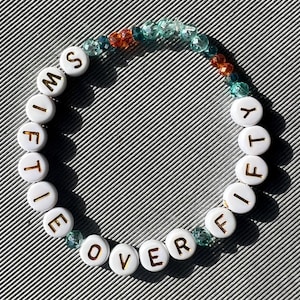 May include: A circular beaded bracelet featuring white round beads with gold lettering that spells out SWIFTY OVER FIFTY. The bracelet is accented with small, translucent, faceted beads in shades of teal, light blue, and orange, arranged in a segment.