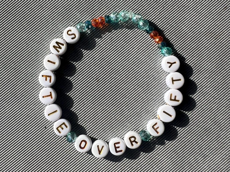May include: A circular beaded bracelet featuring white round beads with gold lettering that spells out SWIFTY OVER FIFTY. The bracelet is accented with small, translucent, faceted beads in shades of teal, light blue, and orange, arranged in a segment.