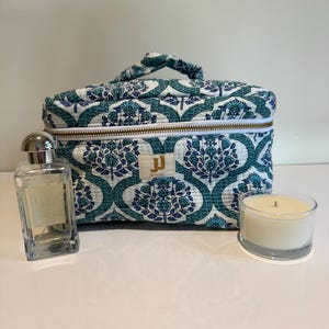 May include: A teal and white patterned cosmetic bag, a perfume bottle, and a candle are arranged. The bag features a floral design. The perfume bottle is rectangular with a silver cap. The candle is in a clear glass.