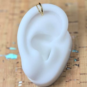 Peut inclure: Un ear cuff doré avec une fine chaîne. Le bijou est présenté sur un modèle d'oreille blanc, sur un fond texturé brun clair. Ce bijou est conçu pour être porté sans perçage.