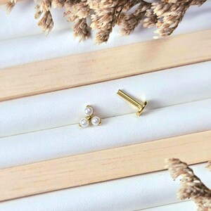 Peut inclure: Bijoux dorés, dont une boucle d'oreille avec trois petites perles blanches en forme triangulaire et une tige unique. Les bijoux sont présentés sur une surface blanche avec des accents en bois et des éléments floraux séchés.