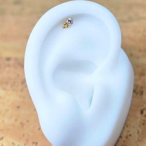 May include: A gold-coloured ear piercing stud featuring a cluster of small gold beads and a single, round, light purple gemstone. The stud is displayed on a white ear-shaped form against a neutral background.