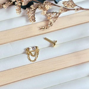 May include: A gold-tone body jewelry stud featuring a rectangular clear gemstone set horizontally. Two delicate gold chains drape from the base of the stone setting. The piece includes a separate flat-back post component for secure attachment.