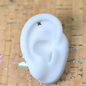 Peut inclure: Une petite boucle d'oreille en forme d'étoile de couleur or est présentée sur un modèle d'oreille blanc. La boucle d'oreille est une étoile à quatre branches. Le fond est une surface texturée brun clair.