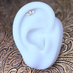 Peut inclure: Un clou d'oreille doré avec trois pierres opalescentes en forme de marquise. Le bijou est posé sur un modèle d'oreille blanc, sur un fond marron texturé.