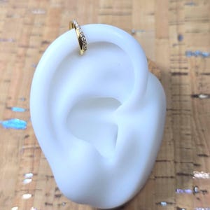 Puede incluir: Un pendiente de oreja dorado con una fila de pequeñas piedras preciosas transparentes. Se muestra en un modelo de oreja blanco sobre un fondo texturizado marrón claro.
