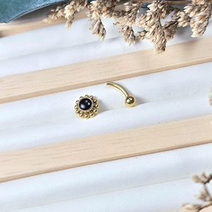 Peut inclure: Un piercing de nombril doré et une petite boucle d'oreille dorée avec une pierre centrale bleu foncé. Les bijoux sont présentés sur une surface blanche avec des accents en bois et des éléments floraux séchés.