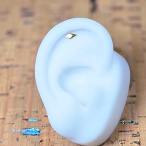 Peut inclure: Un petit clou d'oreille doré est placé dans le haut d'une oreille blanche. Le clou d'oreille a une forme géométrique. Le modèle d'oreille est posé sur une surface texturée brun clair.