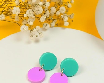 May include: A pair of dangle earrings featuring a teal disc connected to a smaller, pink disc. The earrings are displayed on a white surface with a yellow background and white flowers.