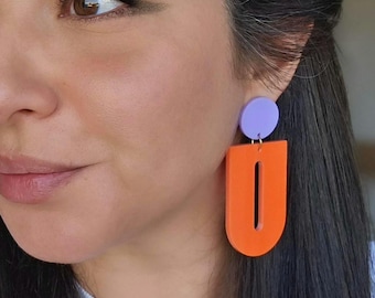 May include: A pair of geometric earrings. Each earring features a small, round, light purple stud at the top, connected to a large, orange, U-shaped pendant with a vertical cutout. The earrings are a bold, modern accessory.