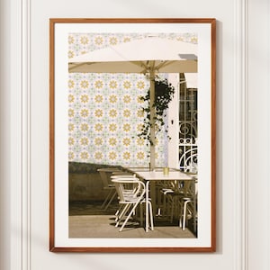 Pode incluir: Uma impressão emoldurada de uma cena de café ao ar livre. A imagem apresenta mesas e cadeiras brancas sob um grande guarda-sol branco. Uma parede com um padrão de azulejos florais repetidos em amarelo, verde e azul serve de fundo. A moldura é castanho quente.