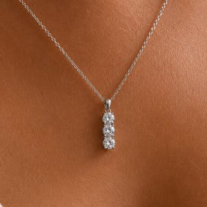 May include: A silver necklace with a pendant of three clear, round gemstones set in a vertical line. The fine chain and pendant create a classic, elegant design. The necklace is shown against a neutral background, highlighting the gemstones.