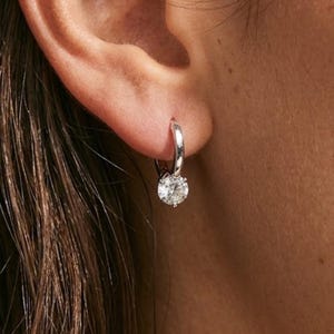 Op de afbeelding: Een enkele zilveren oorring met een ronde, heldere edelsteen. De oorring is bevestigd aan een oor, met de focus op de sieraden. De edelsteen is rond en facetgeslepen en vangt het licht op. Een eenvoudig, elegant ontwerp.