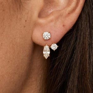 Puede incluir: Primer plano de una oreja adornada con tres pendientes de diamantes diferentes. El pendiente superior es un diamante redondo con talla brillante. Debajo, se ve un diamante con talla marquesa y un diamante redondo más pequeño. Los pendientes están engarzados en oro.