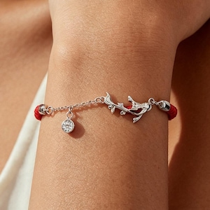 Può includere: Un bracciale in cordino rosso con una catenina in metallo argentato, un piccolo ciondolo circolare con una pietra trasparente e un ornamento centrale in metallo argentato a forma di ramo stilizzato, fissato con terminali in metallo argentato.