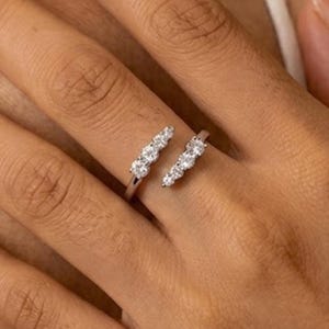 May include: A silver-coloured open ring featuring two distinct ends, each set with a row of three small, clear, round-cut stones. The band is polished and metallic, with the two ends separated by a gap, creating a bypass-style design on the finger.