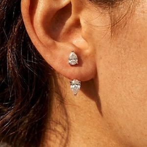 Könnte beinhalten: Nahaufnahme eines Paares tropfenförmiger Diamantohrringe. Die Ohrringe sind in einem hellfarbigen Metall gefasst. Die Diamanten sind klar und funkelnd, und die Ohrringe sind ein klassischer und eleganter Stil, der am Ohr getragen wird.
