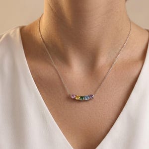 May include: A silver-toned necklace featuring a delicate chain and a curved bar pendant set with five small, round-cut gemstones in a gradient of pink, yellow, green, blue, and purple, resting against a plain white fabric neckline.