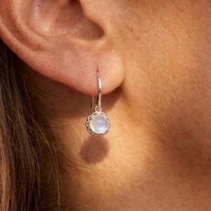 May include: A silver drop earring featuring a round, iridescent moonstone cabochon set within a scalloped metal bezel. The earring hangs from a simple silver hook, displaying a soft, milky white glow with subtle blue light reflections.