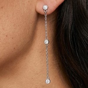 May include: A silver-toned earring with three clear, round gemstones. The top gemstone is attached to the ear, with a delicate chain suspending two additional gemstones below. The earring is shown against a blurred background.