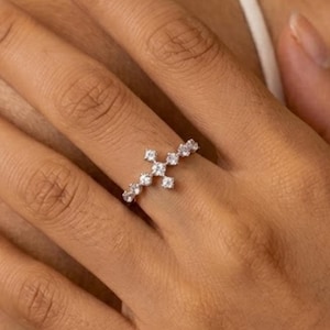 Peut inclure: Une bague argentée ornée d'une croix composée de petites pierres rondes transparentes. L'anneau est fin et métallique, avec le motif en croix centré sur le dessus, mettant en valeur une série de pierres blanches facettées et brillantes.