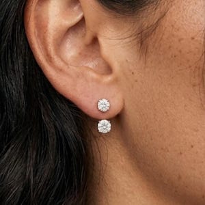 Pode incluir: Close-up de uma orelha com dois brincos de diamante redondos e brilhantes. Os brincos são cravejados em um metal de cor clara e são posicionados um acima do outro no lóbulo da orelha. O tom de pele é castanho claro.