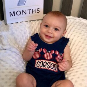 May include: A baby boy wearing a blue and red crab-themed onesie with the name "Carson" embroidered on it. The baby is sitting on a white blanket and smiling at the camera. A white card with the number "7" and the word "MONTHS" is visible in the background.