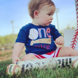 May include: A baby boy wearing a blue shirt with a baseball and the word "one" on it, sitting on a baseball bat. He is holding a baseball in his hand.
