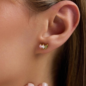 May include: Gold-toned stud earrings featuring three marquise-cut gemstones in pink, white, and green. The earrings are set close together in a curved design, creating a delicate and elegant look. The earrings are shown on an ear.