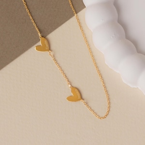 May include: A gold-toned chain necklace featuring two small, flat heart-shaped charms integrated into the chain. Each heart is engraved with a name, specifically Carrie on the upper heart and Jessica on the lower heart, in a simple font.