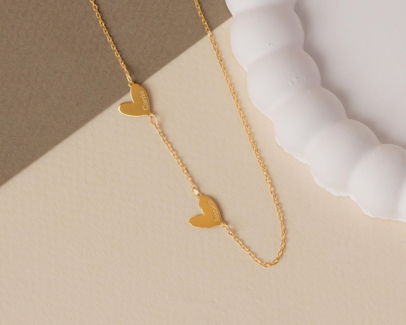 May include: A gold-toned chain necklace featuring two small, flat heart-shaped charms integrated into the chain. Each heart is engraved with a name, specifically Carrie on the upper heart and Jessica on the lower heart, in a simple font.