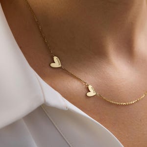May include: A gold-toned necklace featuring a delicate chain with two small, flat heart-shaped charms. Each heart is engraved with a name, specifically Jessica and Carrie, positioned along the length of the chain in a simple, linear arrangement.