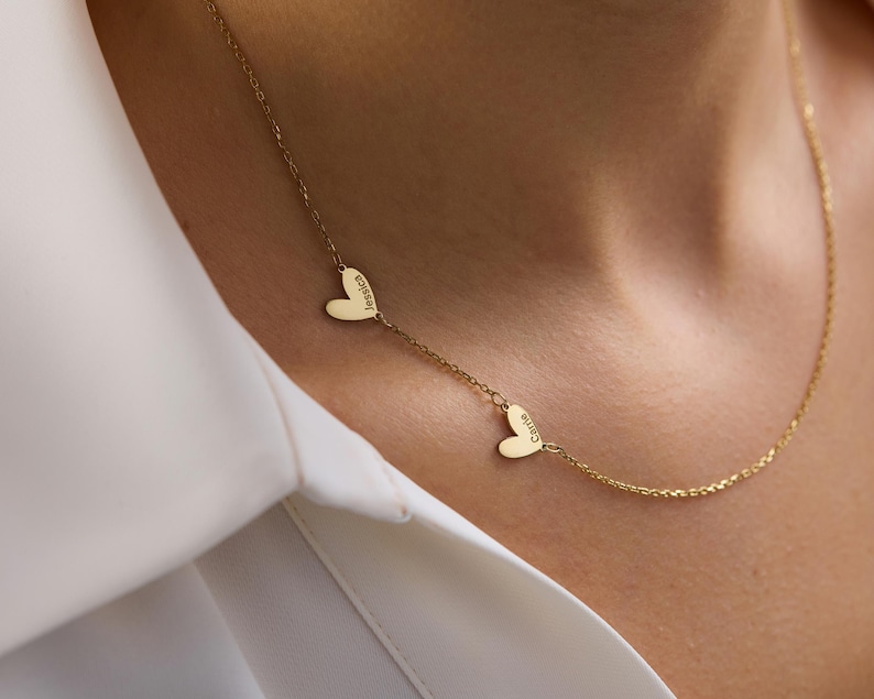 May include: A gold-toned necklace featuring a delicate chain with two small, flat heart-shaped charms. Each heart is engraved with a name, specifically Jessica and Carrie, positioned along the length of the chain in a simple, linear arrangement.