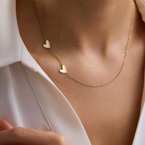 May include: A gold-toned necklace featuring a delicate chain and two small heart-shaped charms. Each heart charm is engraved with a name, specifically Jessica and Carrie, positioned asymmetrically along the length of the thin metal chain.