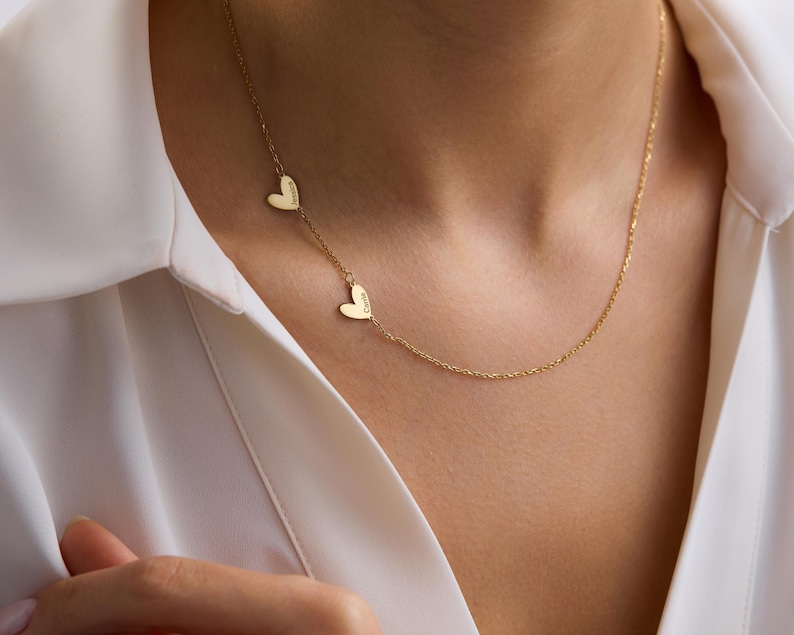 May include: A gold-toned necklace featuring a delicate chain and two small heart-shaped charms. Each heart charm is engraved with a name, specifically Jessica and Carrie, positioned asymmetrically along the length of the thin metal chain.