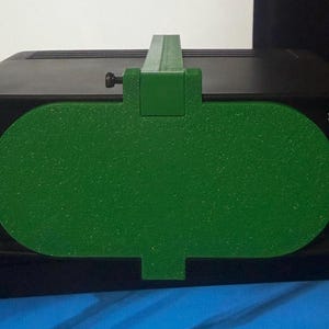 Puede incluir: Un dispositivo electrónico negro con un accesorio verde. La pieza verde tiene una superficie texturizada y una forma rectangular con extremos redondeados. Se ve un tornillo en el lateral. El texto "Elgato AF Lens Timer" es visible.