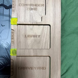 Könnte beinhalten: Kartenorganizer aus Holz mit lasergraviertem Text. Der Organizer hat Fächer mit den Aufschriften "COMMANDER ZONE", "LIBRARY" und "GRAVEYARD". Die Kanten haben neongrüne Akzente. Für Kartenspiele.
