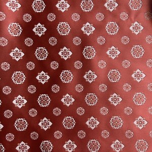 May include: A close-up of a fabric featuring a repeating floral design. The base colour is a deep reddish-brown, with small, stylised flowers in white and light brown. The flowers are arranged in a scattered, all-over pattern.
